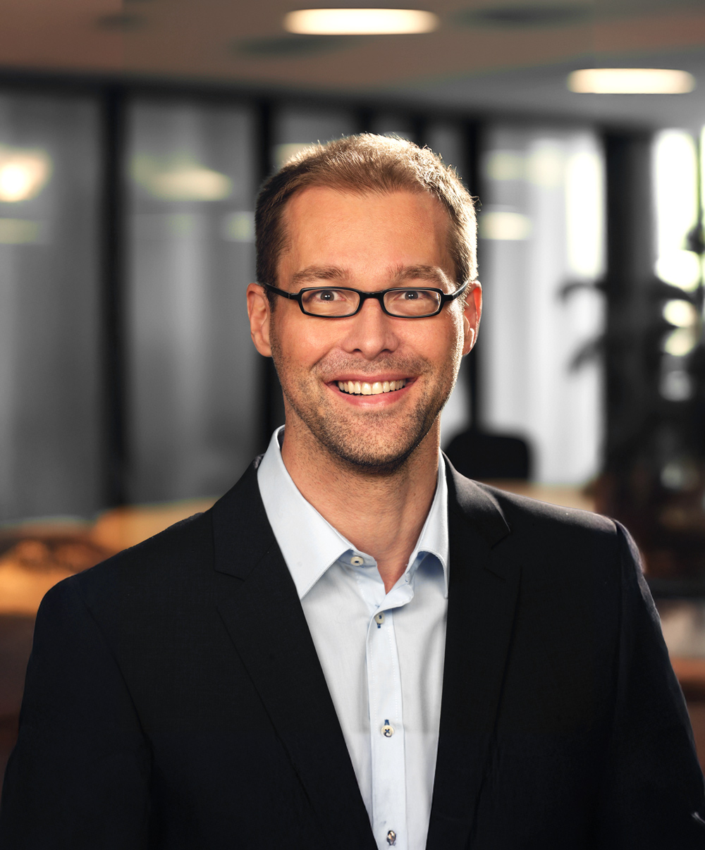 Detlef Bruns, Inhaber von Bruns Consulting IT Service GmbH, mit Brille und Anzug in einem modernen Büro, freundlich lächelnd.
